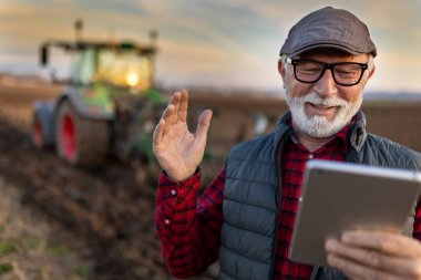 Excited mature farmer working on tablet in front of tractor in field
