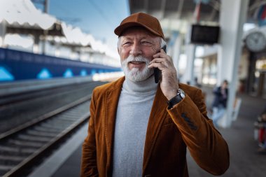 Happy senior man talking on mobile phone while waiting train on terminal at railway station