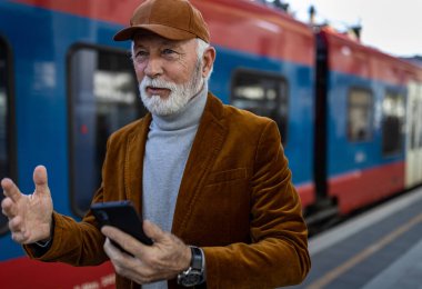Senior man using mobile phone in front of train on terminal at station