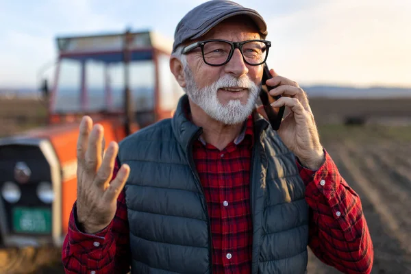 Excited mature farmer talking on mobile phone in front of tractor in field