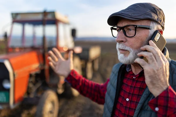 Furious mature farmer talking on mobile phone in front of tractor in field