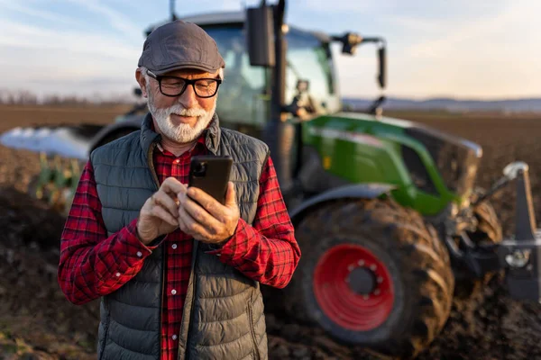 Smiling mature farmer looking at mobile phone in front of tractor in field. Smart farming concept