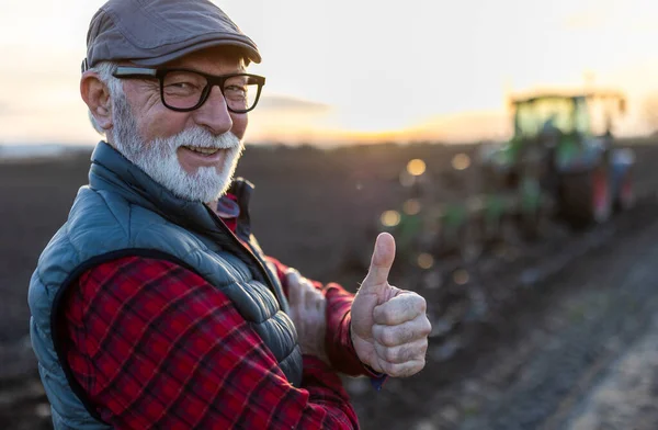 Portrait of mature farmer showing thumb up in front of tractor in field in autumn