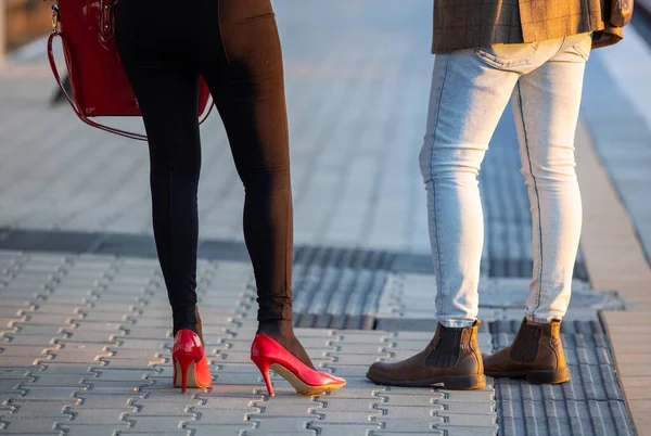 Close up of couple's legs standing on paving outdoors, woman in high red heels and man in stylish shoes