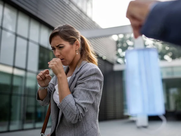 Man offering protective mask to business woman while coughing in front of office building. Virus protection and health care responsibility concept