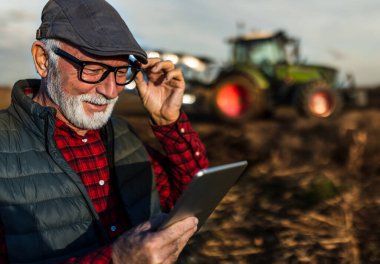 Excited mature farmer working on tablet and thinking about business plans in front of tractor in field