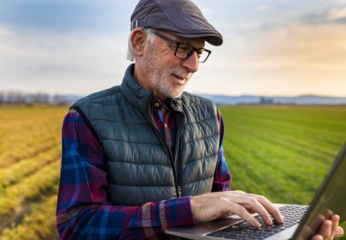 Satisfied senior farmer working on laptop in field in early spring time