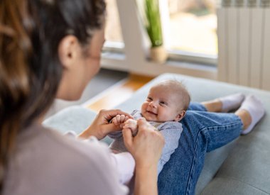 Genç anne evdeki kanepede oturuyor ve kucağında güzel bir bebek, kalçasında gülüyor ve oynuyor. Annelik aşkı, ilgi ve neşe konsepti