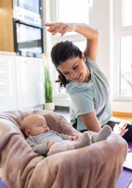 Genç ve güzel anne evde mutfak dolaplarının önünde yoga minderi üzerinde egzersiz yapıyor ve yeni doğmuş bebeğinin uyumasını seyrediyor. Sağlıklı zihinsel ve fiziksel durum kavramı