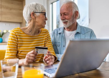 Mutfak arka planında yemek masasında oturan ve kredi kartıyla internetten alışveriş yapan neşeli yaşlı çift. 