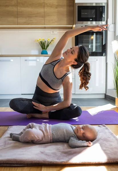 年轻漂亮的母亲在厨房橱柜前的瑜伽垫上锻炼 看着她刚出生的孩子在睡觉 劳动后的身心健康概念 — 图库照片