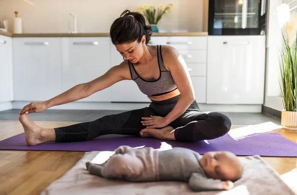 年轻漂亮的母亲在厨房橱柜前锻炼 在家里用瑜伽垫伸展腿 而新生儿则睡在旁边 劳动后的身心健康概念 — 图库照片