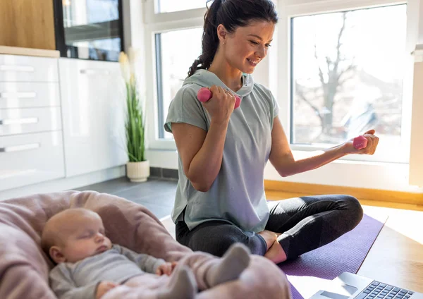 年轻漂亮的妈妈看着笔记本电脑 在家里用哑铃在瑜伽垫上锻炼 而她的新生婴儿就睡在她身边 劳动后的身心健康概念 — 图库照片