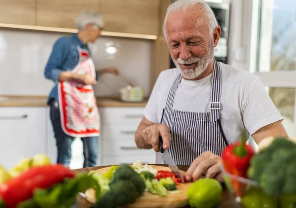 シニアカップルはスープを 夫は野菜を切り 妻は料理を背景に — ストック写真