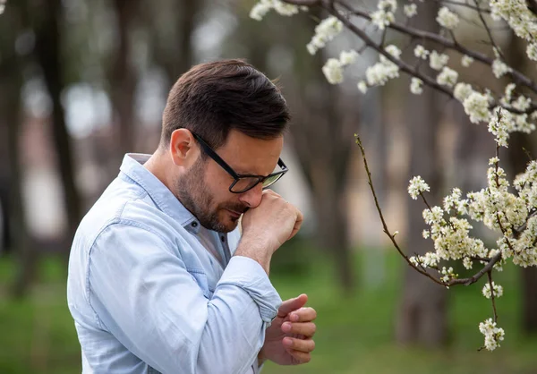 Handsome man having nose itching and sneezing beside blooming tree in spring outdoors. Allergy pollen symptoms concept