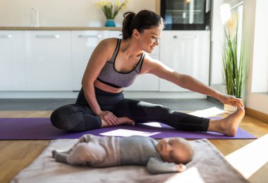 Yeni doğmuş bir bebek yanında uyurken evde mutfak dolaplarının önünde yoga minderi üzerinde egzersiz yapan genç ve güzel bir anne. Sağlıklı zihinsel ve fiziksel durum kavramı
