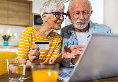 Mutfak arka planında yemek masasında oturan ve kredi kartıyla internetten alışveriş yapan neşeli yaşlı çift. 