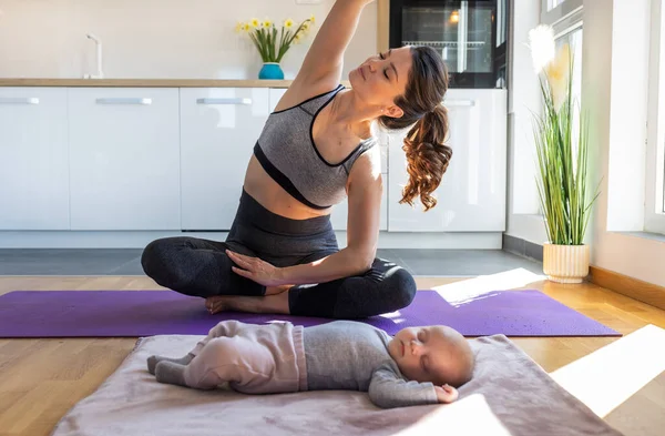 年轻漂亮的母亲在厨房橱柜前的瑜伽垫上锻炼 而新生儿则在睡觉 劳动后的身心健康概念 — 图库照片