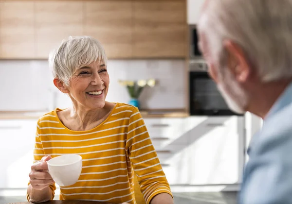 笑着的老夫妇坐在厨房橱柜前的餐桌前 喝咖啡 在家里聊天 — 图库照片