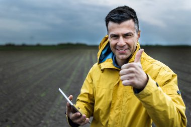 Rüzgarlı kış gününde tarlada dikilen ve baş parmağını gösteren tabletli çiftçi.