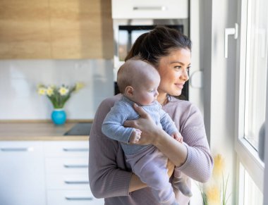 Gülümseyen güzel anne kucağında bebek ve evde pencerenin yanında hassas anın tadını çıkarıyor. Annelik aşkı, ilgi ve neşe konsepti