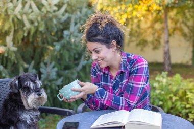 Pretty young woman sitting in garden with dog on chair, reading book and drinking tea clipart