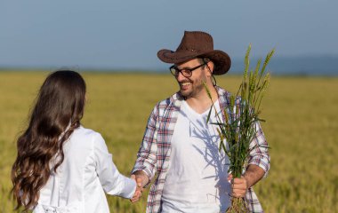 Beyaz önlüklü çiftçi ve kadın tarım uzmanı yazın buğday tarlasında durup el sıkışıyorlar.