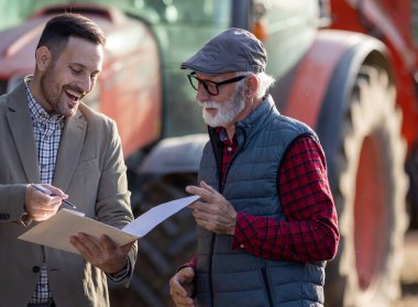 Çiftlikteki traktör ve tarım makinelerinin önünde işadamıyla sözleşme imzalayan kıdemli çiftçi.
