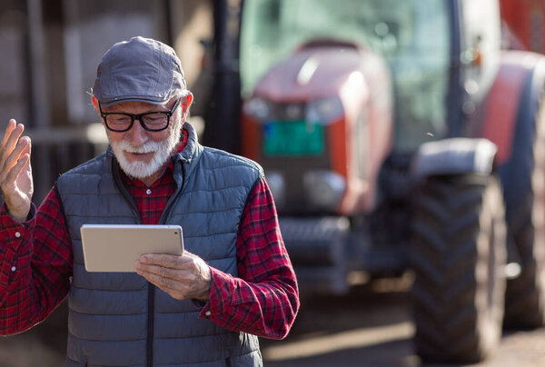 Portrait of excited senior man looking at tablet in front of tractor in cowshed on cattle ranch