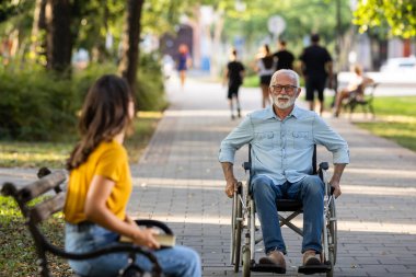 Yaşlı adam tekerlekli sandalye kullanırken genç kadın bankta oturuyor ve parkta bekliyor.  