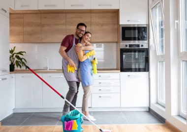 Genç çift, önlük giyip ellerinde sprey şişelerle modern mutfaklarını temizledikten sonra sarılıp eğleniyorlar.