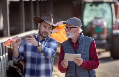 İnek ahırının ve traktörün önünde konuşan ve tablet kullanan iki gülen çiftçi.