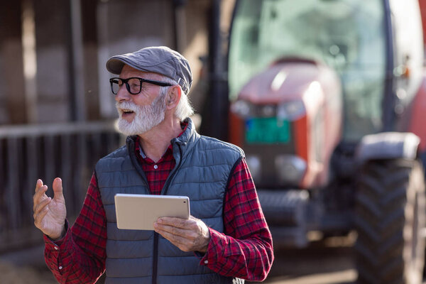 Portrait of excited senior man holding tablet in front of tractor in cowshed on cattle ranch