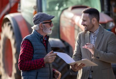 Gülümseyen iş adamı ve kıdemli çiftçi çiftlikte traktörün önünde bankayla kredi sözleşmesi hakkında konuşuyorlar.
