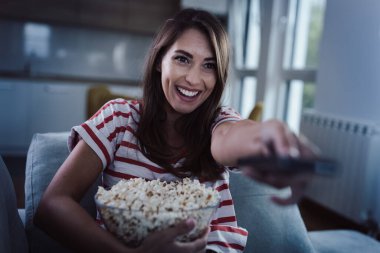 Gülümseyen genç bir kadın elinde patlamış mısır ve kumandayla evinde film seyrediyor.