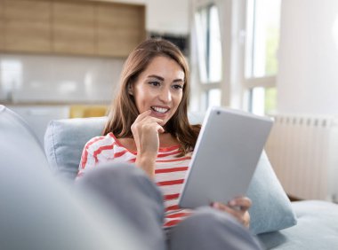 Güzel genç bir kadın rahat bir oturma odasında kanepede oturuyor ve tablete bakıyor.