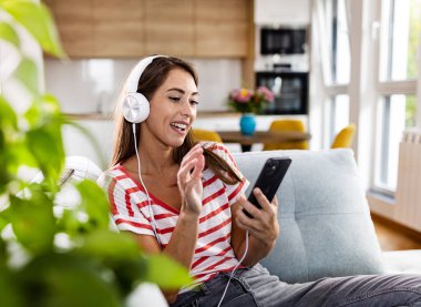 Genç, güzel bir kadın rahat bir ev ortamında kanepede oturuyor, kulaklık takıyor, cep telefonu tutuyor, müzik dinliyor ya da video görüşmesi yapıyor, gülümsüyor, eğleniyor.