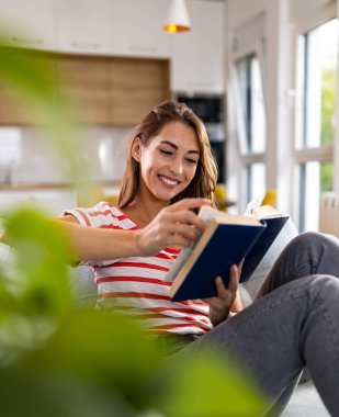 Oturma odasındaki kanepede oturan, arka planda mutfağı olan ve kitap okuyan güzel genç bir kadın.