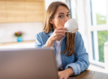Kahve içen, yemek masasında dizüstü bilgisayarda çalışan, arka planda mutfağı olan güzel bir kadın. Ev kavramından çalışmak