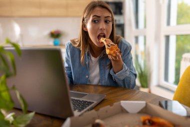 Evde, mutfağın önünde dizüstü bilgisayarda çalışan ve aynı zamanda pizza yiyen meşgul genç bir kadın.