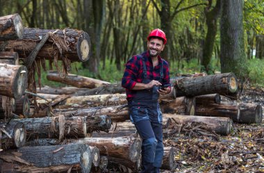 Ormanda istiflenmiş ağaç gövdelerinin yanında cep telefonu tutan yakışıklı orman mühendisi.