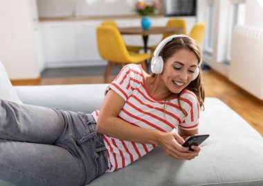 Genç ve güzel bir kadın rahat bir ev ortamında kanepede uzanıyor, kulaklık takıyor ve cep telefonu tutuyor, müzik dinliyor ya da video görüşmesi yapıyor, gülümsüyor, eğleniyor.