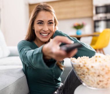 Gülümseyen genç bir kadın elinde patlamış mısır ve kumandayla evinde film seyrediyor.