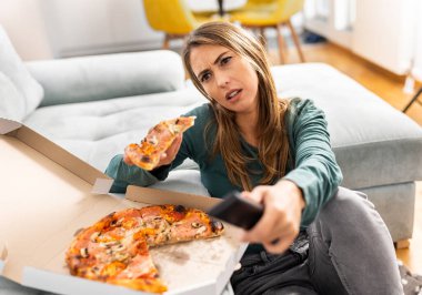 Güzel genç bir kadın oturma odasındaki koltuğun yanında pizza yerken uzaktan kumandayla en sevdiği TV programını izliyor.