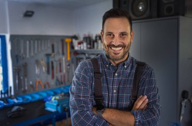 Atölyede kolları çapraz duran yakışıklı bir tamirci portresi, arka planda duvarda asılı aletler.