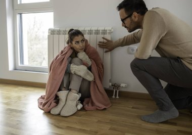 Genç bir kadın battaniyeyle kaplı radyatörün yanında otururken, bir adam dairede ısıtıcının sıcaklığını kontrol ediyor.