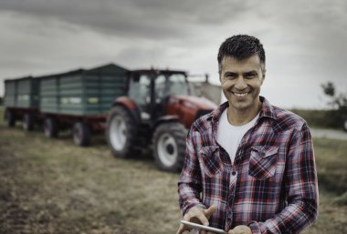 Sonbaharda hasattan sonra arka planda iki römorkla birlikte traktörün önünde tablet tutan yakışıklı çiftçinin portresi.