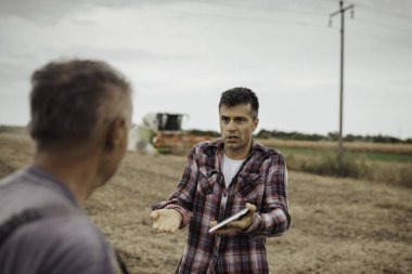 Soya tarlasında dikilen ve diğer çiftçilerle konuşan üzgün çiftçi, sonbaharda arka planda çalışan hasat makinesini birleştiriyor.