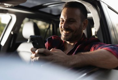Ekose gömlekli yakışıklı adam araba sürüyor ve cep telefonuyla mesajlaşıyor.