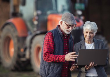Çiftlikte konuşan ve traktörün önünde dizüstü bilgisayara bakan kıdemli çiftçi ve olgun iş kadını.
